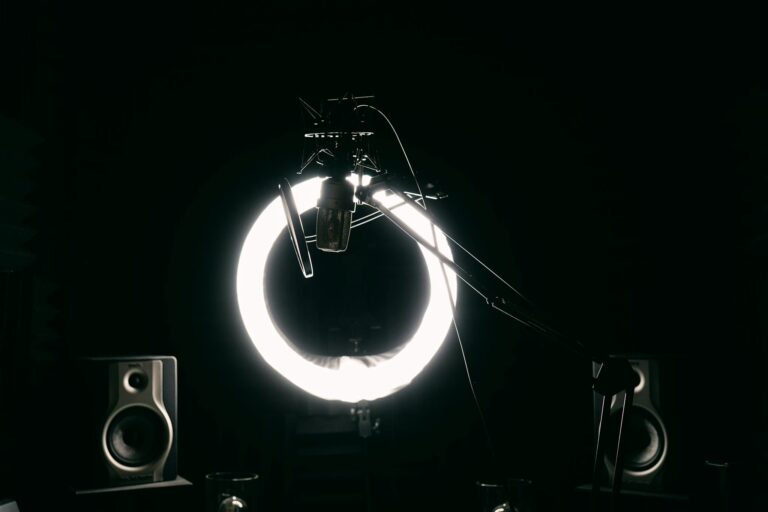 Dark studio room with condenser microphone, ring light, and speakers for recording purposes.